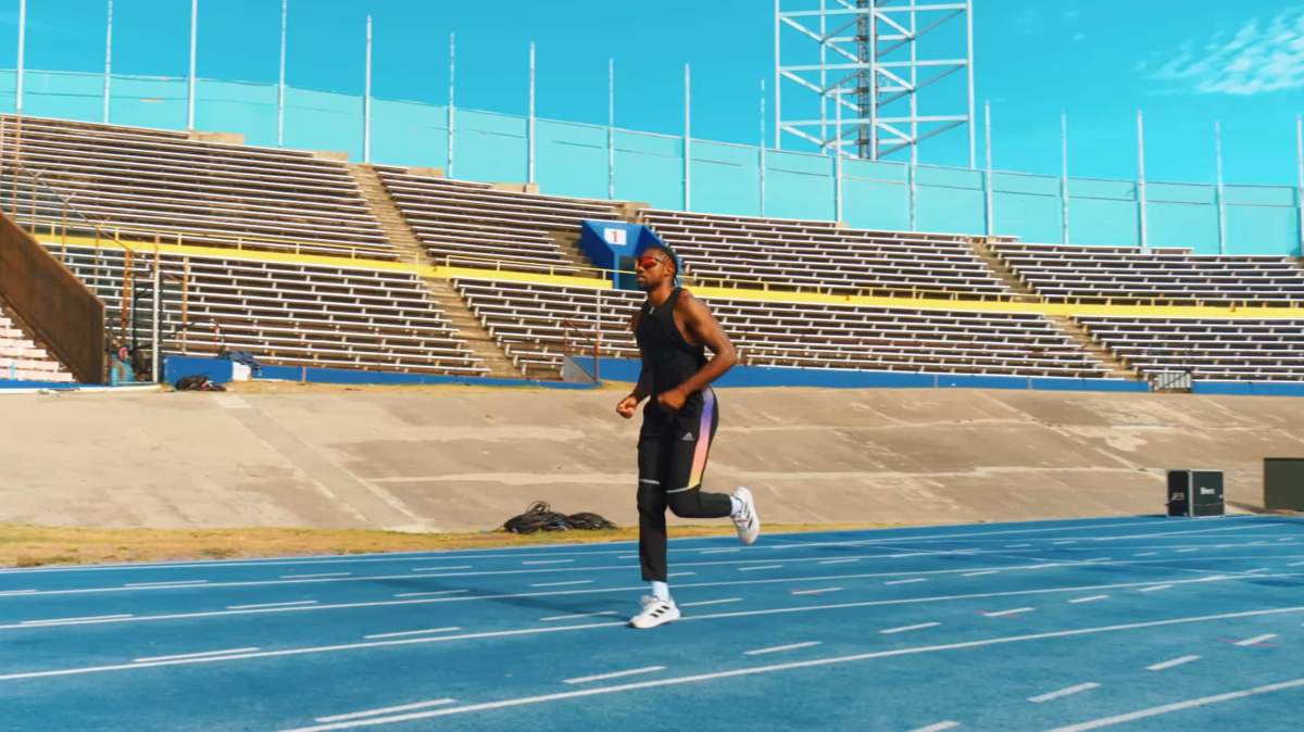 Noah Lyles runs on a track in an empty stadium with sunglasses on.